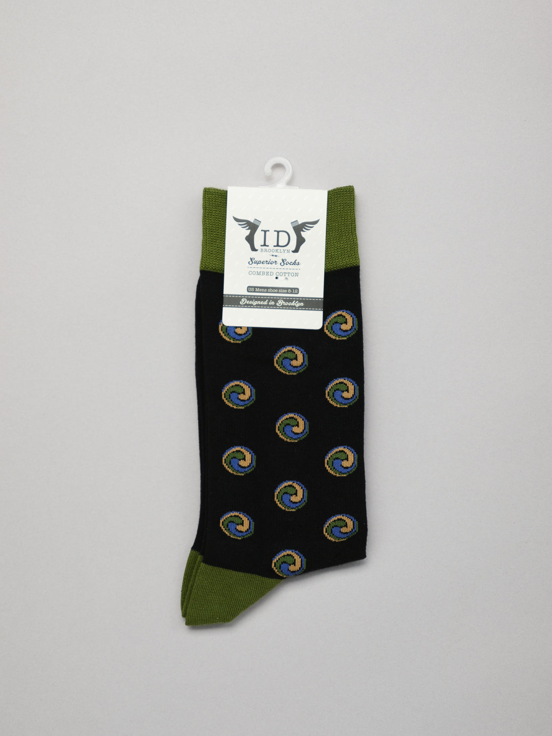 A solitary black Trinity Graphic Sock by ID Menswear features green cuffs and toes in soft combed cotton, with a circular multicolored swirl Jacquard design. A white label with text and design is attached at the top against a plain light gray background.