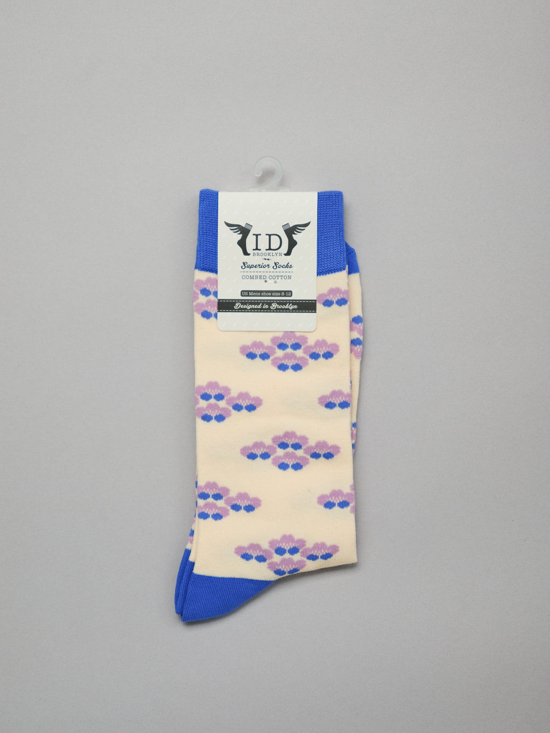 The Yuki Graphic Socks by ID Menswear feature a stylish cream design with whimsical purple, pink, and blue clouds. They have blue cuffs, heels, and toes on a gray background and come with a tag labeled ID adorned with decorative wings.