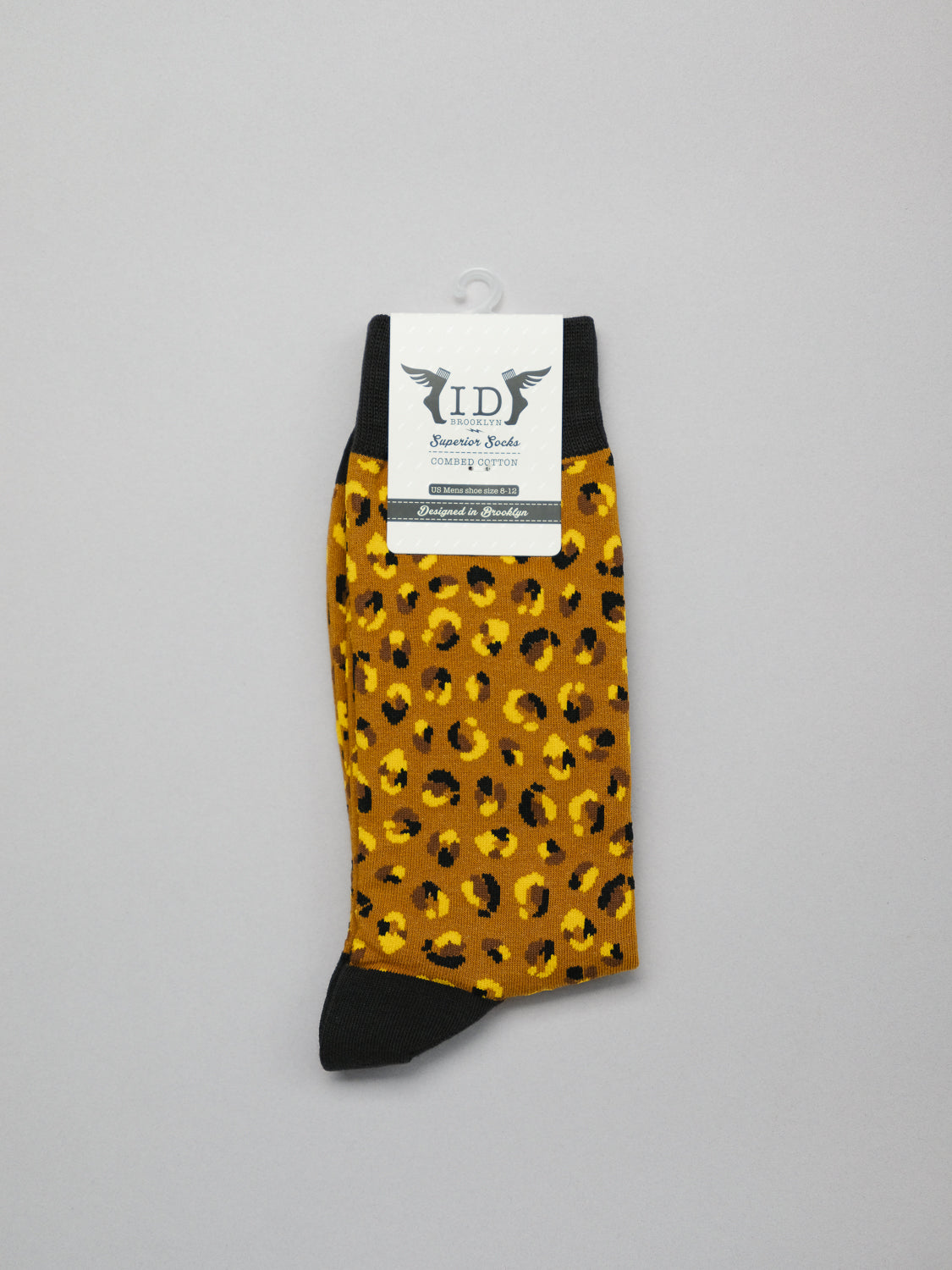 The Cheetah Graphic Sock by ID Menswear features a leopard-print design with a black toe and cuff. Made from soft combed cotton, it includes a white label with black text and an emblem on top, set against a plain background.