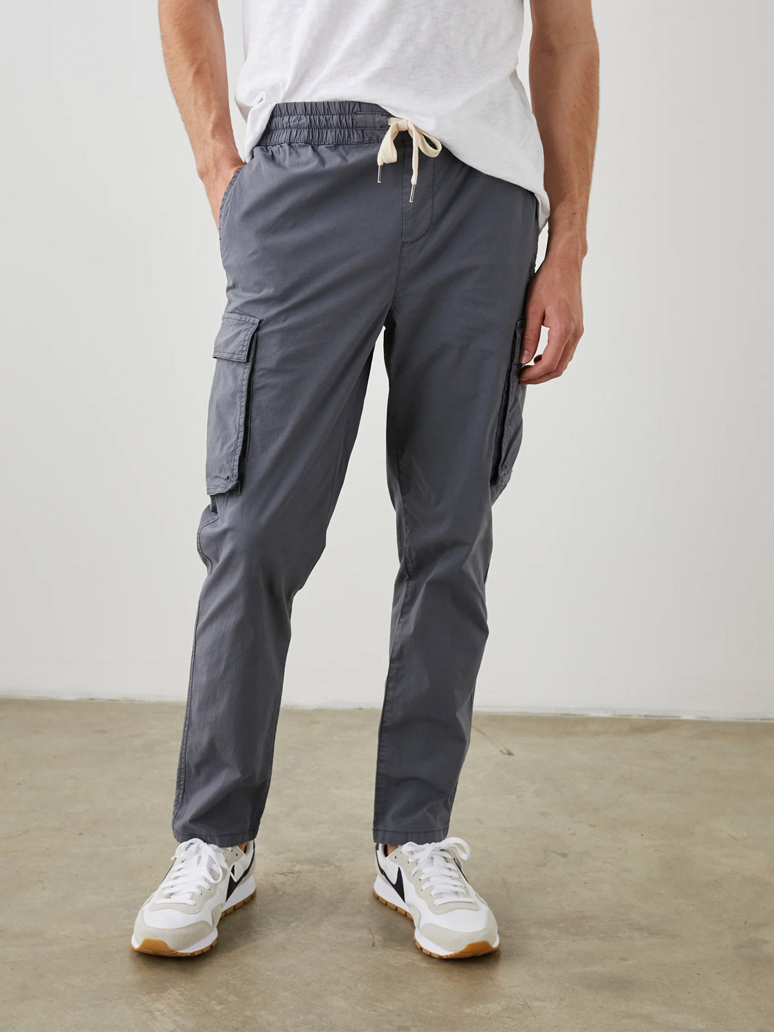A person is wearing a white T-shirt, the Rails Emmerson Pant—a lightweight gray cotton jogger with side pockets and drawstring waist—and white sneakers. The background features a plain light-colored wall and concrete floor.