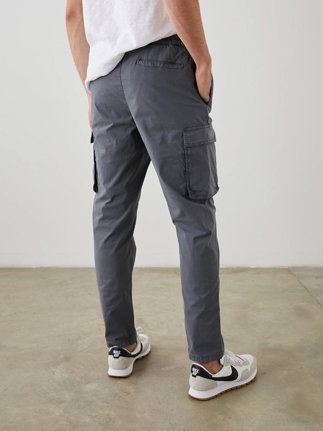 A person in Rails Emmerson Pants—modern gray workwear with handy side pockets, paired with white sneakers and partially visible white shirt—stands on a concrete floor, facing away from the camera.