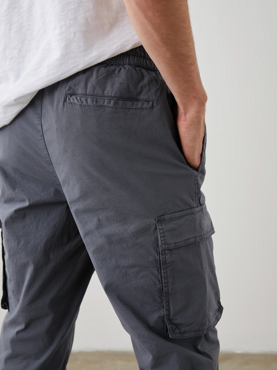 A person wearing a white shirt and the Rails Emmerson Pant in dark gray, made from lightweight cotton, has their left hand in the pocket. These modern workwear pants by Rails feature back and side pockets against a plain indoor background.