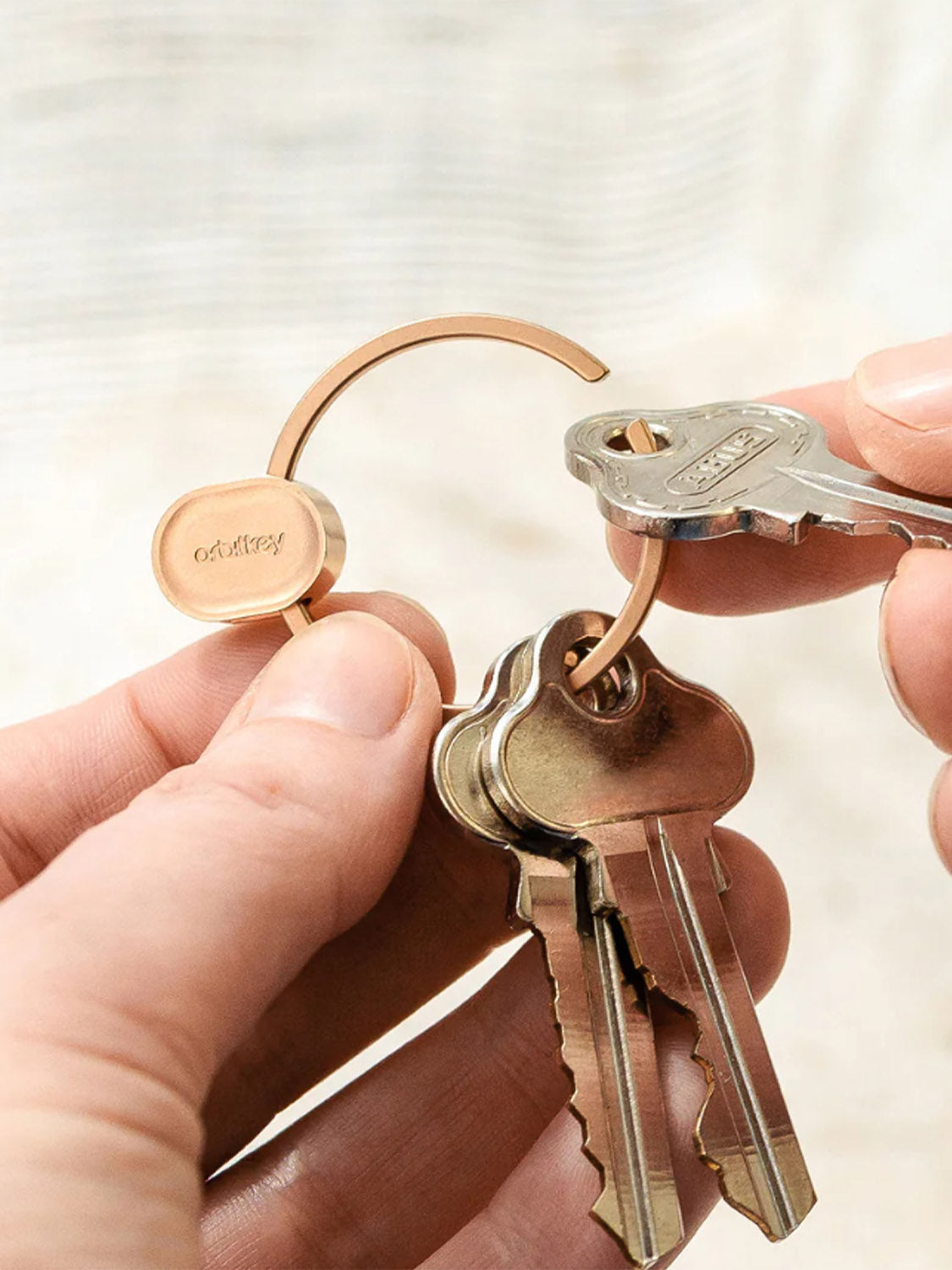 A person's hands attach a silver key to the Orbitkeys Key Ring V2, a durable stainless steel ring already holding several gold-colored keys, with the softly blurred background highlighting the product.