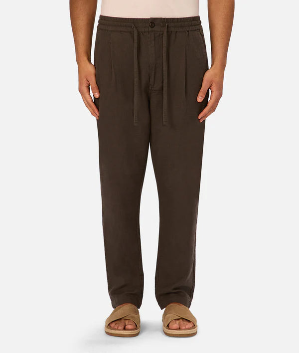 A model wears The Mallorca Pant by Industrie—relaxed-fit, breathable cotton with a drawstring waist in dark brown—paired with tan open-toe sandals, standing against a plain white background.