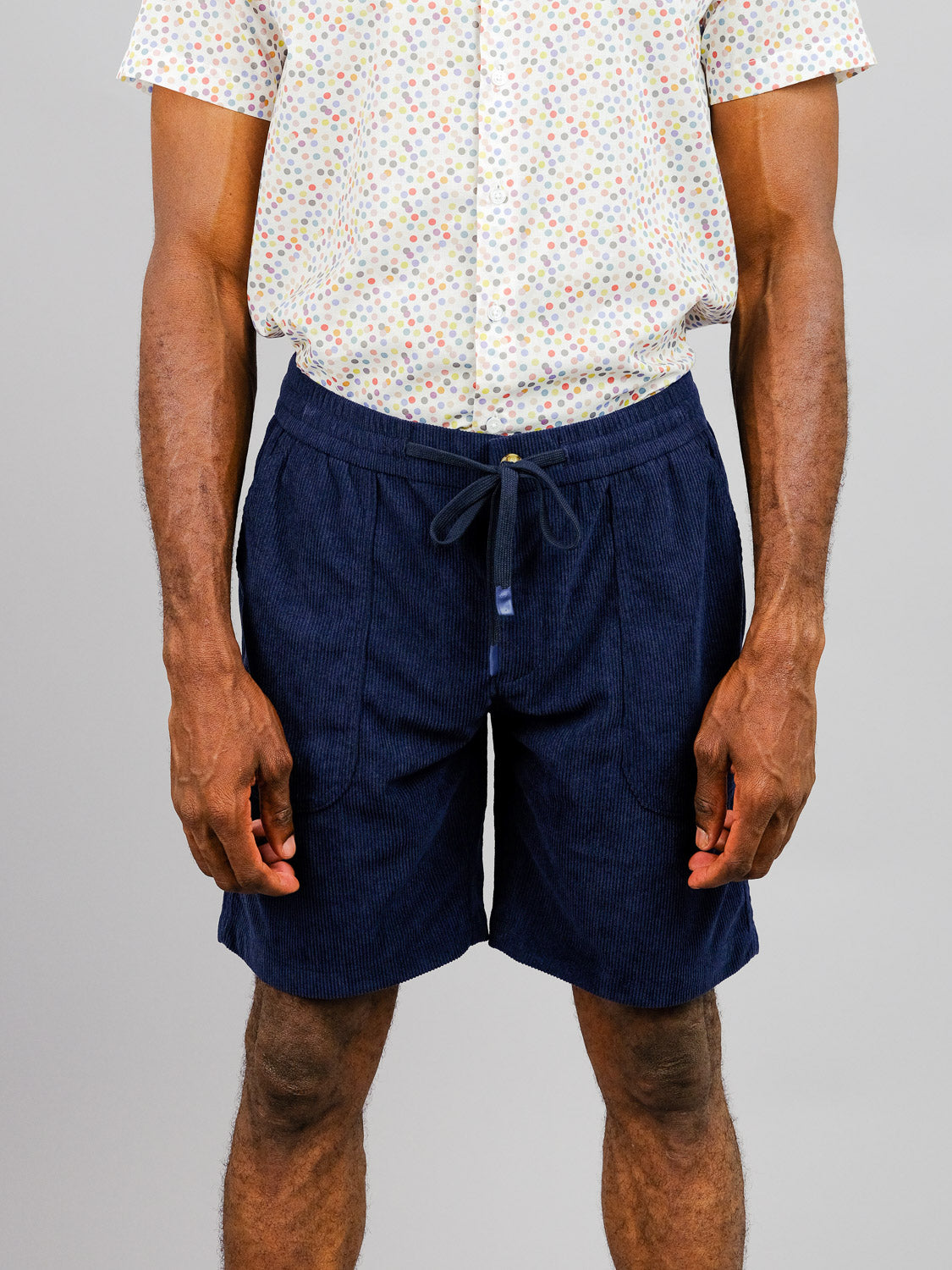A person in a white shirt with colorful polka dots and Tunis Corduroy Shorts by ID Menswear, which feature an elastic waistband, stands against a plain background. The image captures the outfit from mid-torso to knees, ideal for a casual stroll.