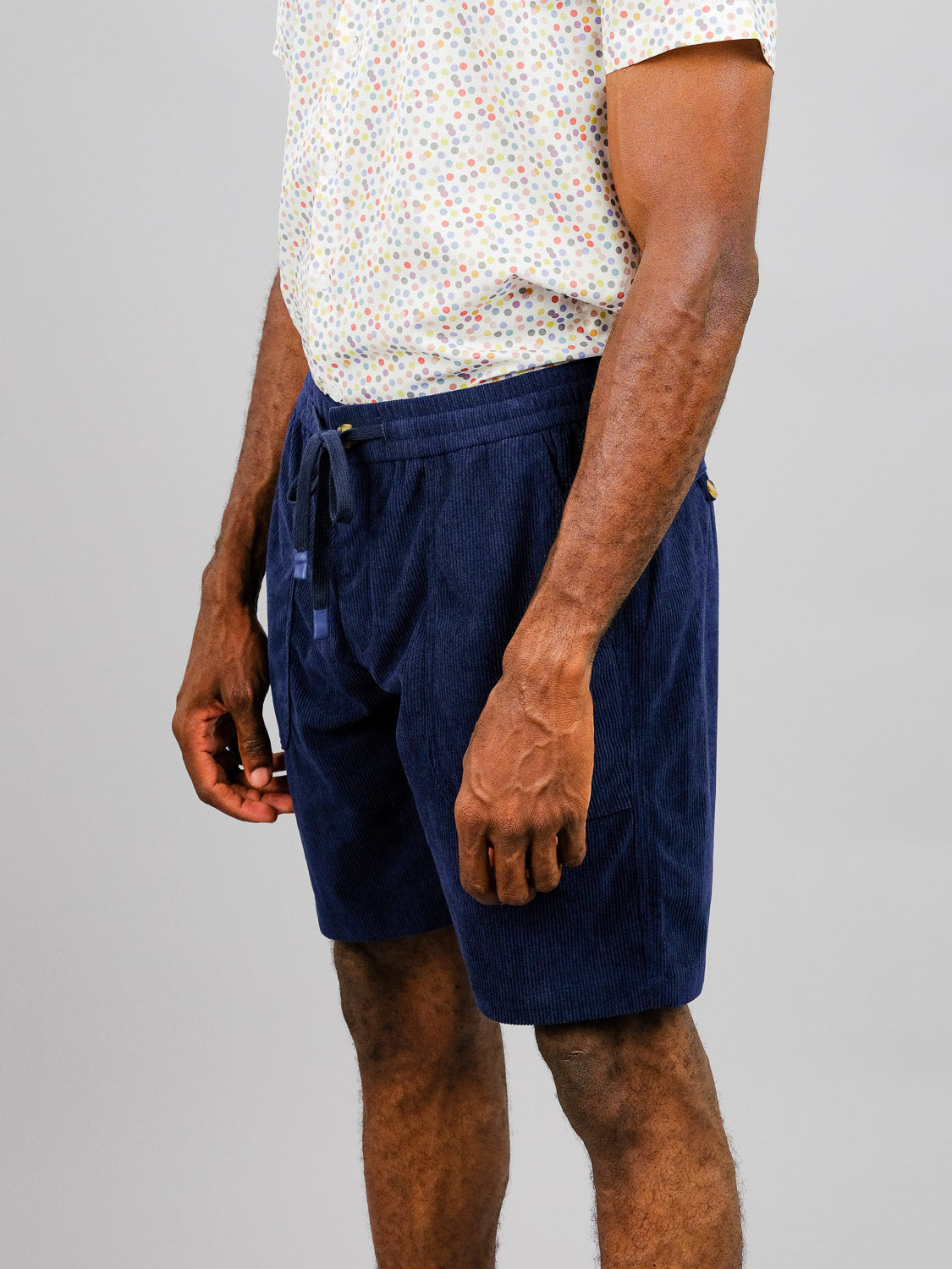 A person casually stands against a neutral background wearing a white shirt with colorful polka dots and a pair of ID Menswears Tunis Corduroy Shorts, which are blue and feature an elastic waistband.