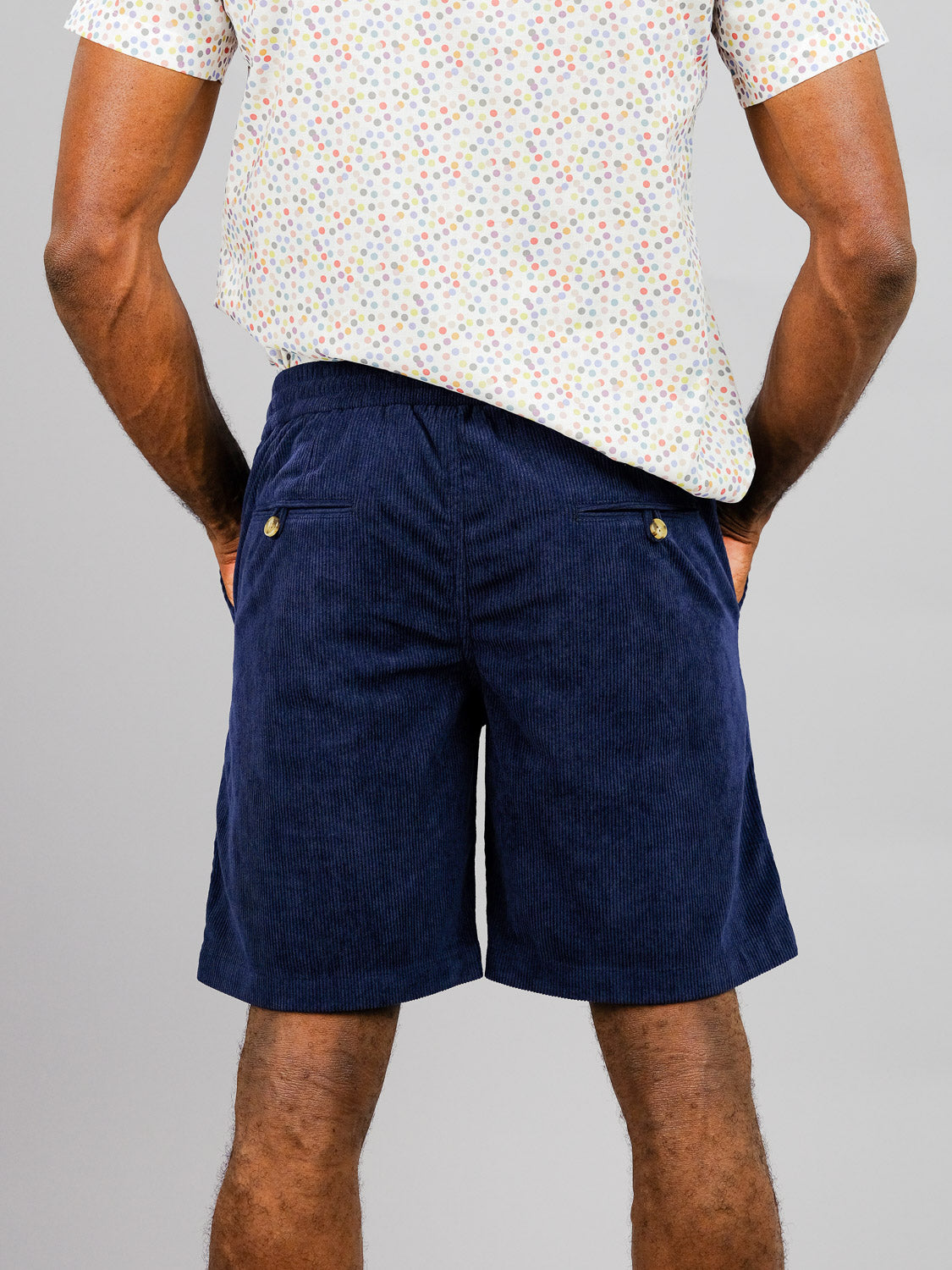 A person wears a white shirt with colorful dots and navy blue Tunis Corduroy Shorts from ID Menswear. The shorts feature an elastic waistband, pockets, and are perfect for a casual stroll against the plain gray background.