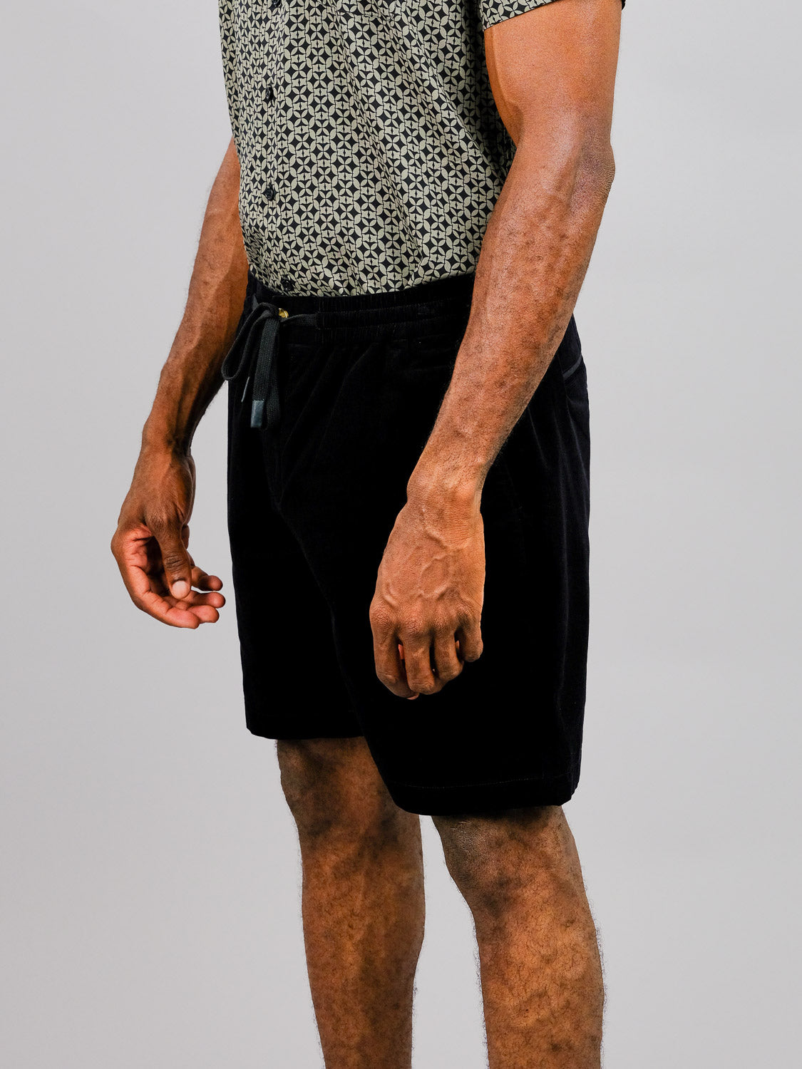 Against a plain background, a person showcases ID Menswears Odyssey Corduroy Stretch Shorts paired with a patterned short-sleeve shirt. The photo highlights the relaxed fit of the ensemble, focusing on the lower torso, arms, and legs.