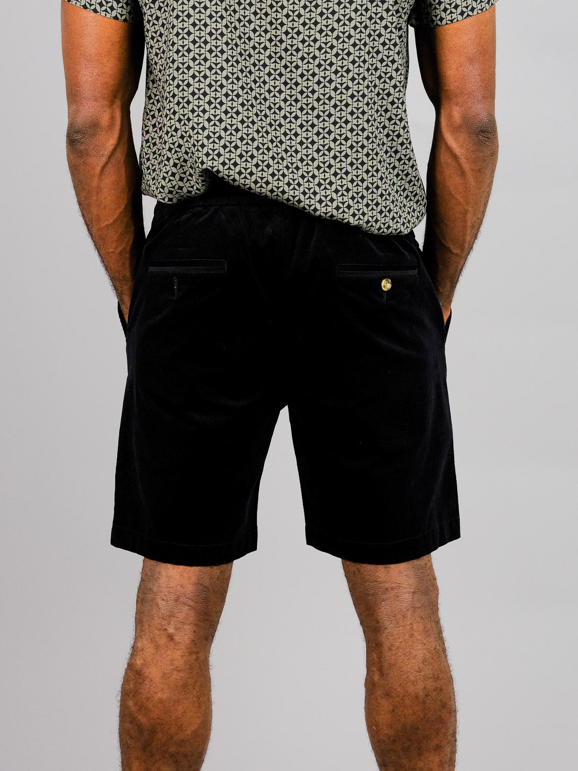 A person stands with their back to the camera, wearing a patterned short-sleeve shirt and ID Menswears Odyssey Corduroy Stretch Shorts. Their hands rest comfortably in their pockets against a plain gray background.