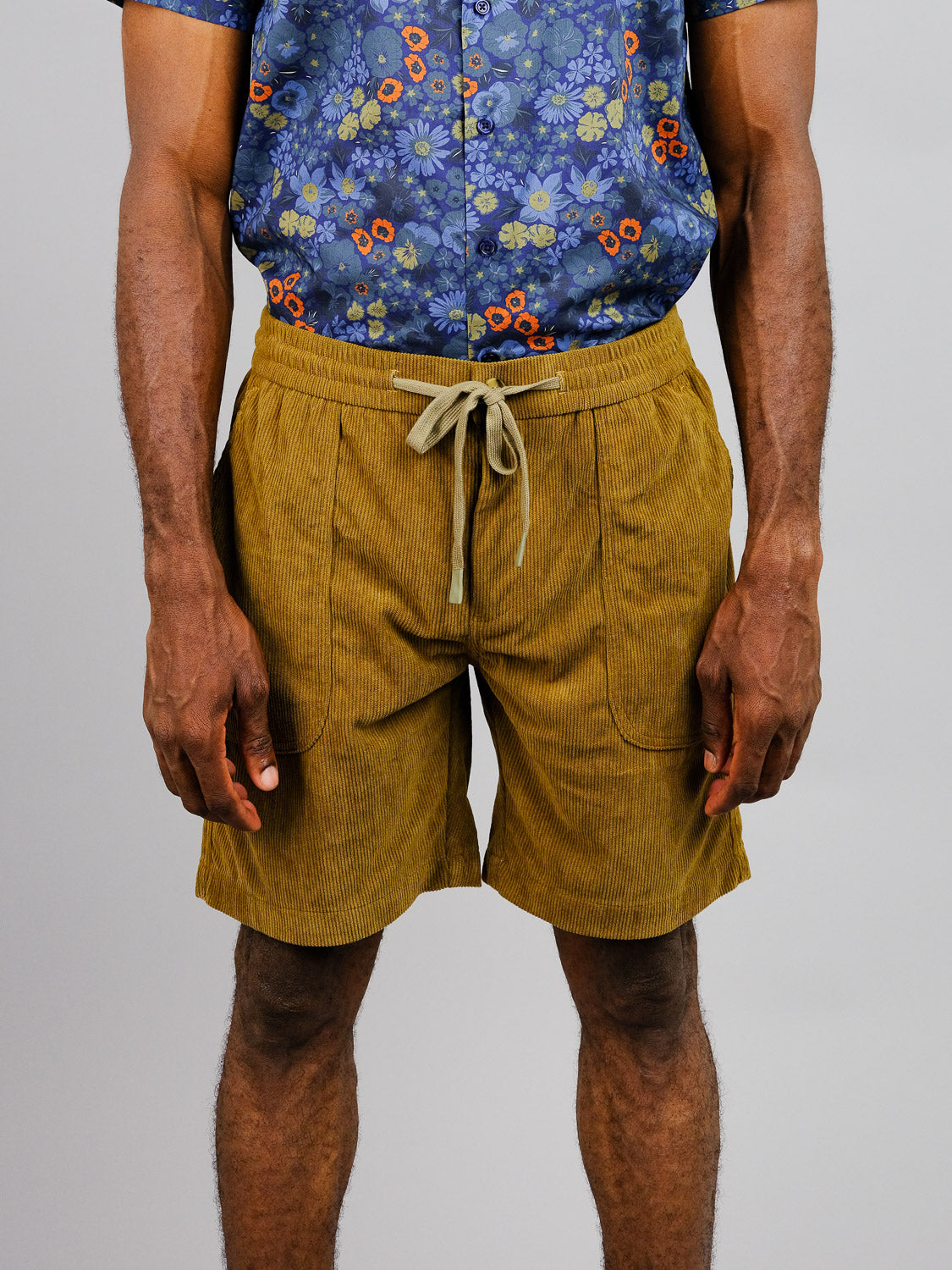 A person stands against a plain backdrop, relaxed and ready for a casual stroll, wearing mustard Tunis Corduroy Shorts by ID Menswear with an elastic waistband paired with a blue floral-patterned shirt.