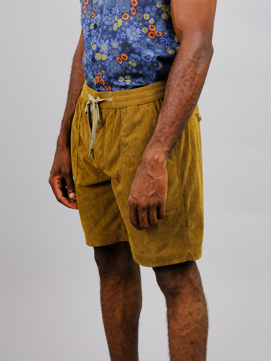 The image features someone in a blue floral shirt and ID Menswears mustard yellow Tunis Corduroy Shorts with an elastic waistband, capturing a casual stroll mood against a plain backdrop from the neck down.