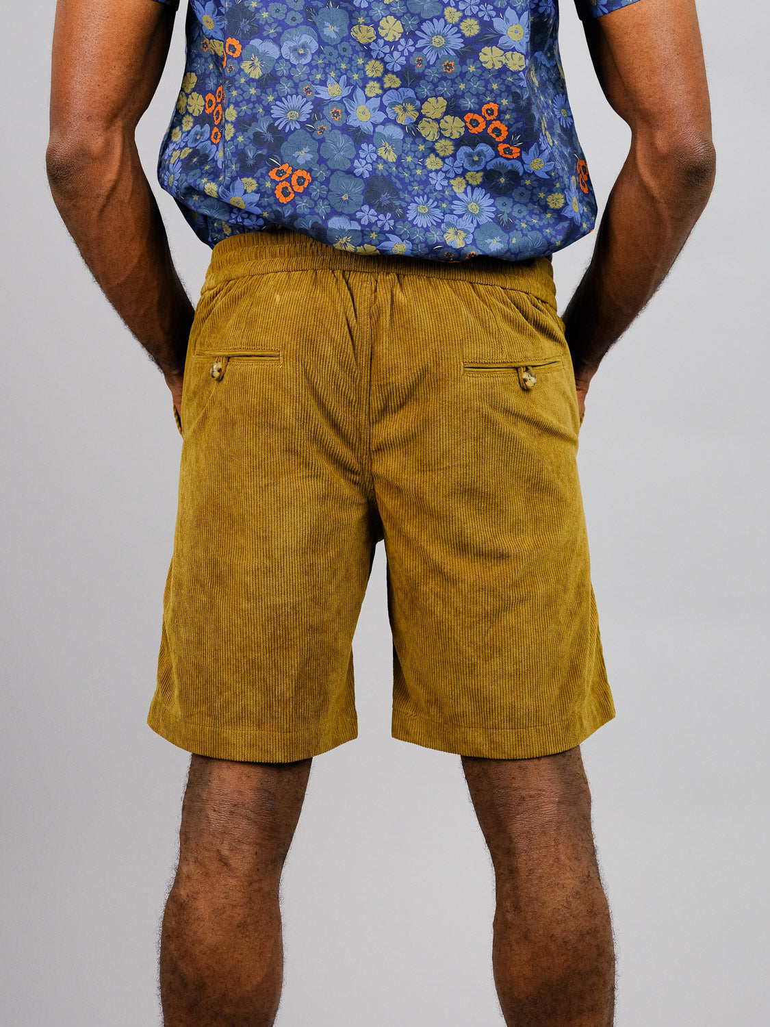 A person in a blue floral shirt and mustard yellow Tunis Corduroy Shorts by ID Menswear, featuring an elastic waistband, stands against a plain gray background. The scene enhances a laid-back vibe, ideal for casual walks.