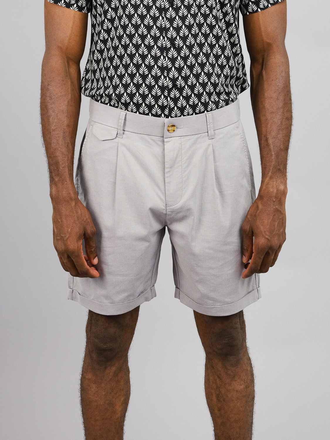 A person is dressed in a black and white patterned shirt and light gray Masai Stretch Shorts from ID Menswear, known for casual comfort. The photo captures the torso and legs with relaxed arms, set against a plain light gray background, enhancing the outfits simplicity.