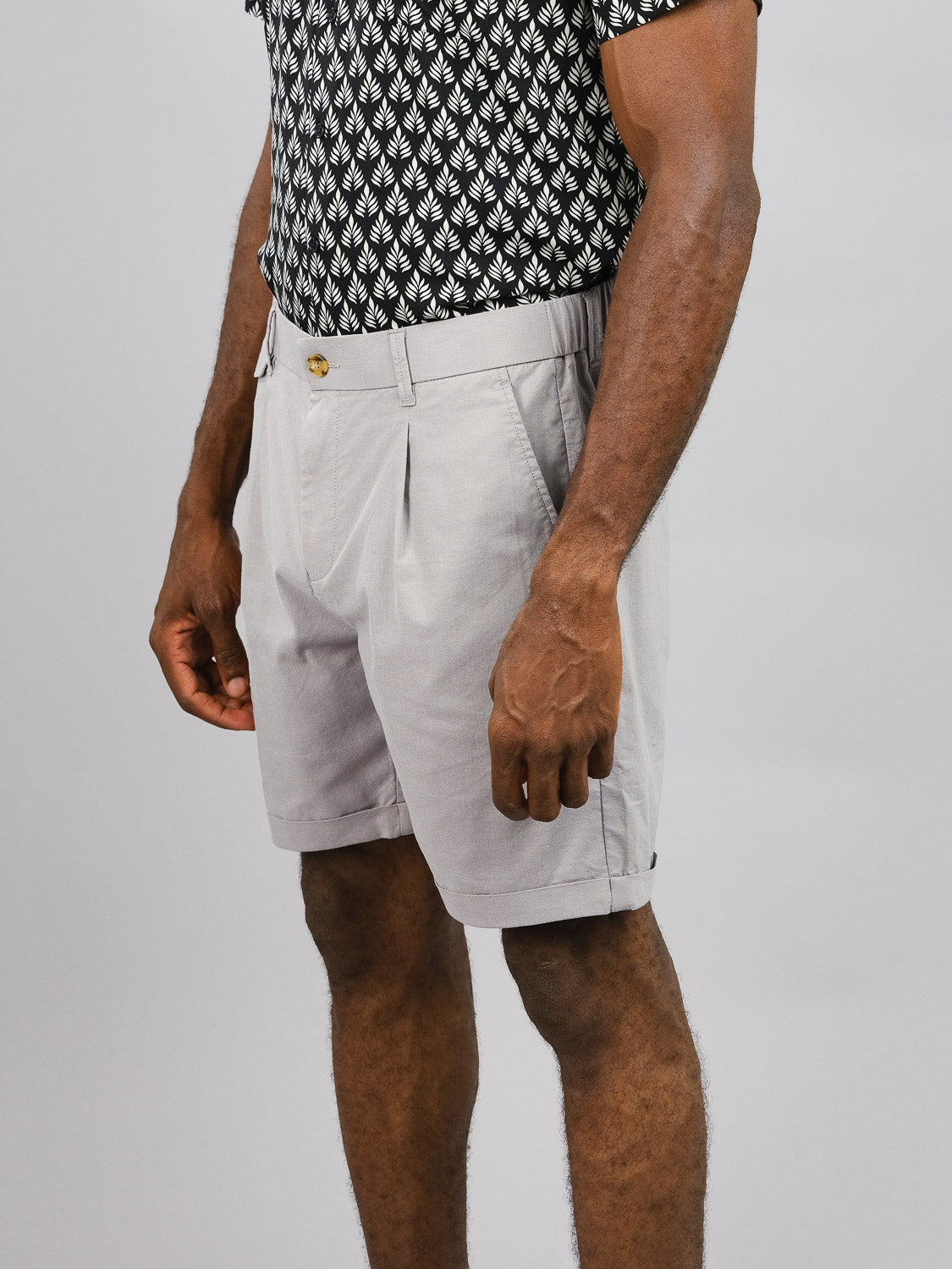 A person stands against a plain background wearing light gray Masai Stretch Shorts from ID Menswear, featuring an elastic waistband and button detail, paired with a short-sleeve shirt adorned with a black and white leaf pattern that perfectly complements the mid-thigh casual comfort of the shorts.