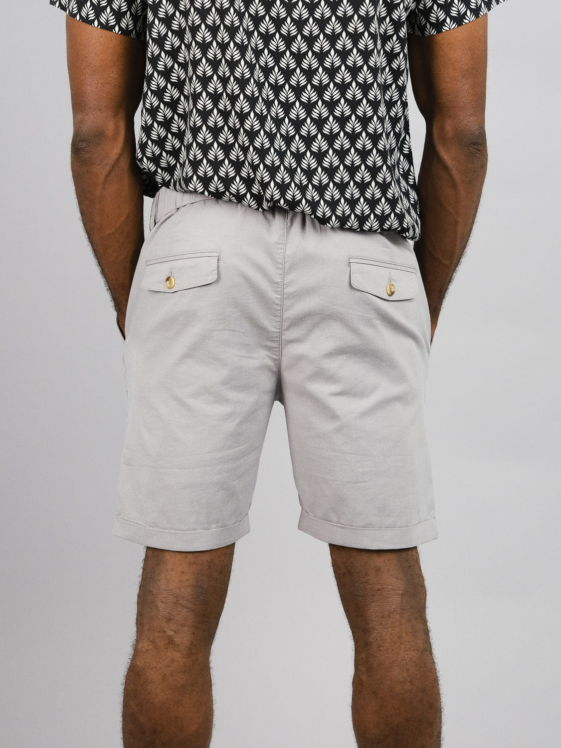 A person is seen from the back wearing a black and white patterned shirt and light gray Masai Stretch Shorts by ID Menswear, which have an elastic waistband and buttoned back pockets, portraying relaxed casualness against a plain gray backdrop.
