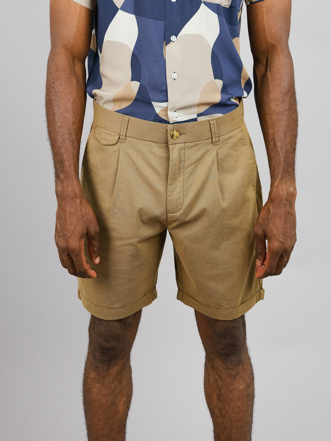 A person wears a patterned shirt with beige, blue, and white shapes paired with tan ID Menswears Masai Stretch Short featuring an elastic waistband for casual comfort, standing against a plain gray background.