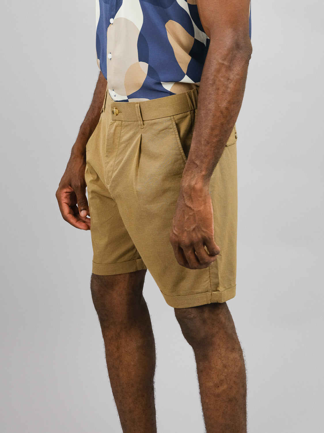 A person wears a blue and beige patterned short-sleeve shirt paired with ID Menswears Masai Stretch Short in tan, featuring an elastic waistband. Their right arm is slightly bent while the left rests by their side, exuding casual comfort against a plain grey background.