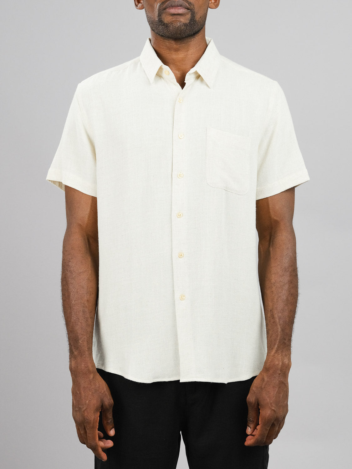 A man in a short-sleeved, light beige New Hampton Linen Blend Shirt from ID Menswear stands against a plain gray background. Made of breathable linen-viscose, it features a single breast pocket. He pairs it with dark pants, arms resting casually by his sides, highlighting the shirt’s details.