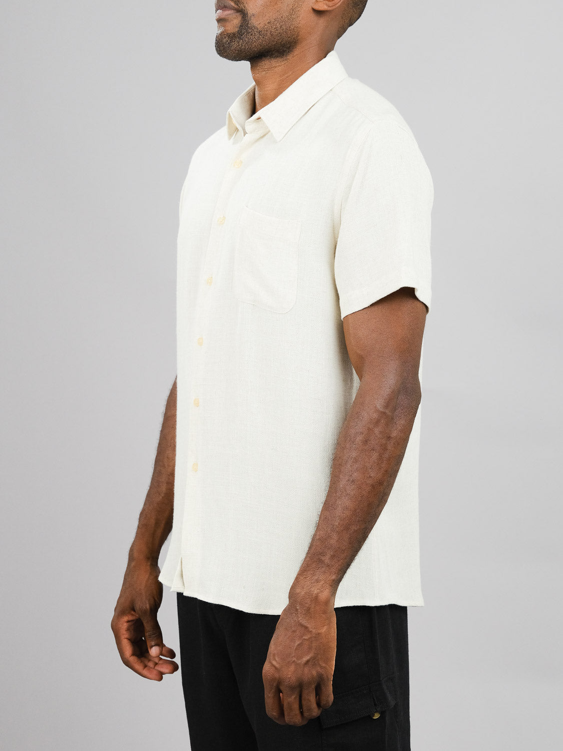 Against a gray backdrop, an individual wears ID Menswears New Hampton Linen Blend Shirt in light beige. The regular-fit, breathable shirt features a chest pocket and is paired effortlessly with black pants as they face slightly left with relaxed arms.