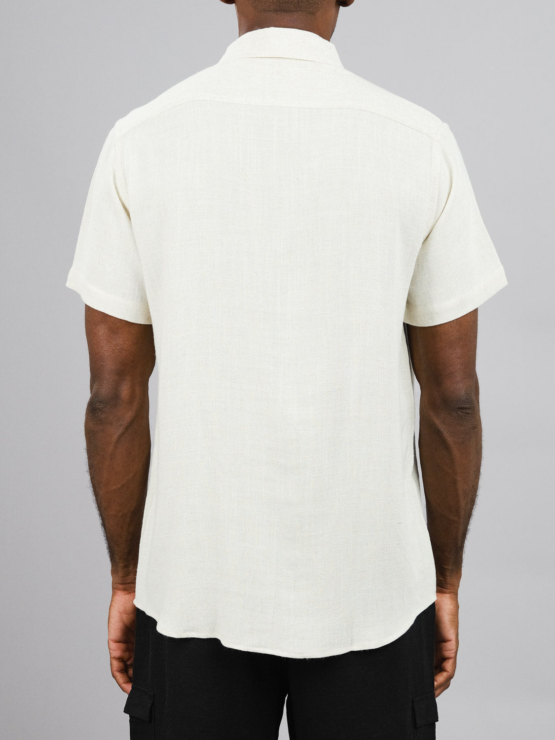 A person is seen from behind wearing a short sleeve, light cream New Hampton Linen Blend Shirt by ID Menswear. The regular-fit, linen-viscose blend shirt is paired with black shorts against a plain gray background.