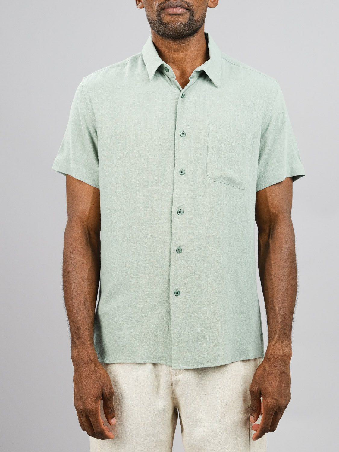 A person is wearing the New Hampton Linen Blend Shirt by ID Menswear, featuring a breathable light green fabric, regular fit, short sleeves, and a left-side pocket. This comfortable linen-viscose blend pairs perfectly with light-colored pants against the plain gray background.