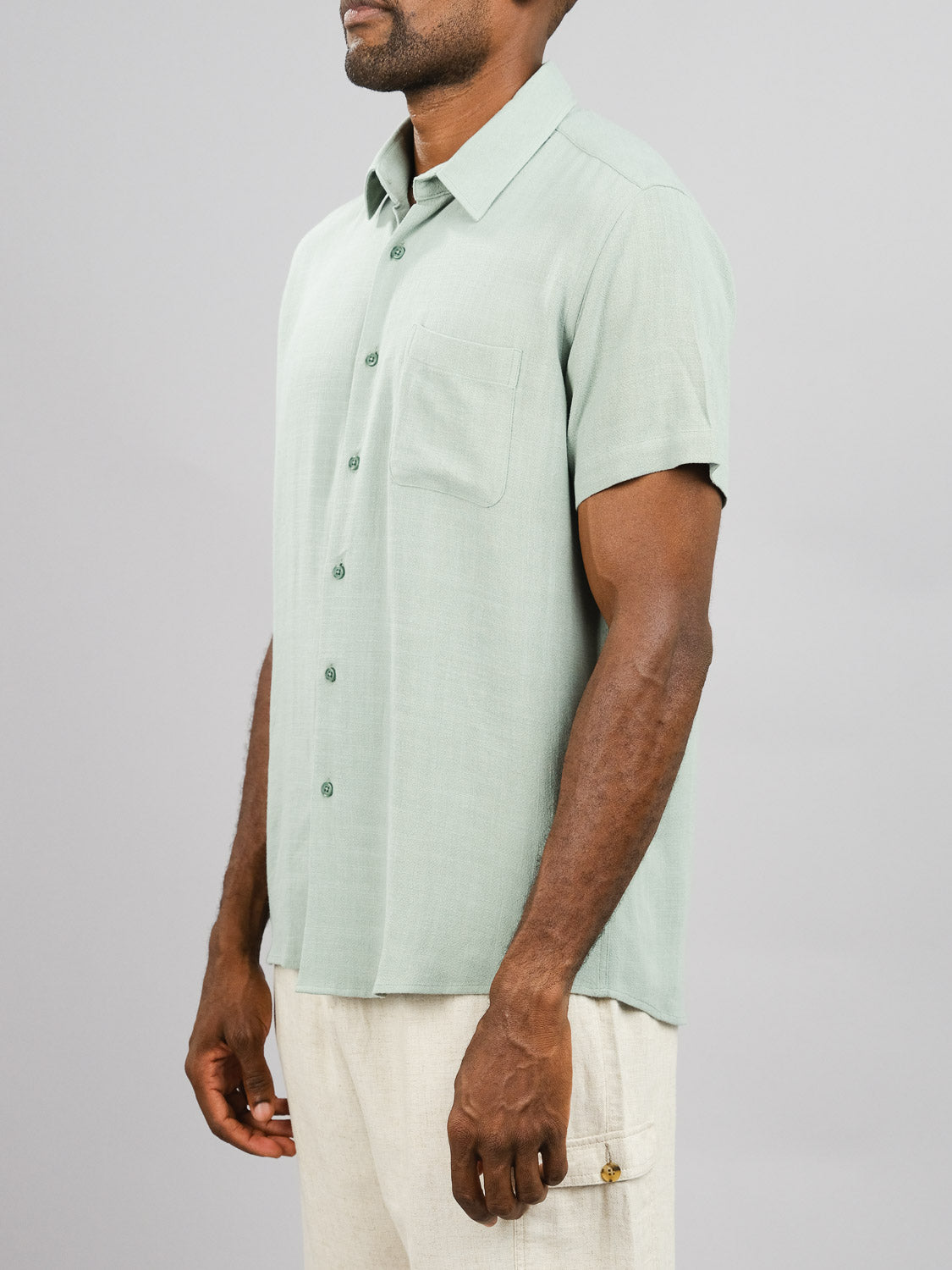 A person wears the New Hampton Linen Blend Shirt by ID Menswear, a light green regular-fit shirt with green buttons and a chest pocket, crafted from a breathable linen-viscose blend. They stand against a plain gray background wearing beige pants and are positioned at a slight angle.