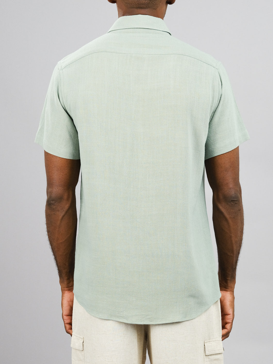 The image features a person from behind wearing an untucked ID Menswears New Hampton Linen Blend Shirt in light green, paired with beige pants, on a plain gray background. The breathable short-sleeve button-up shirt is made of a relaxed linen-viscose blend.