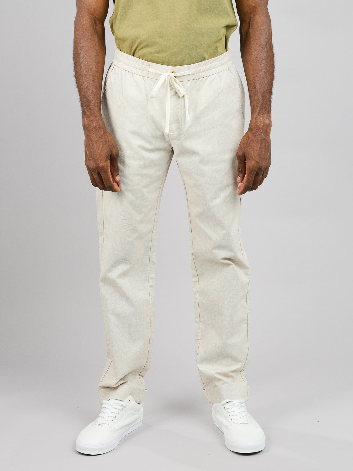 A person stands against a gray background wearing a green shirt, Cruise Lightweight Stretch Pant by ID Menswear, and white sneakers. The shot focuses from the shoulders down, showcasing the lightweight and comfortable nature of the outfit.