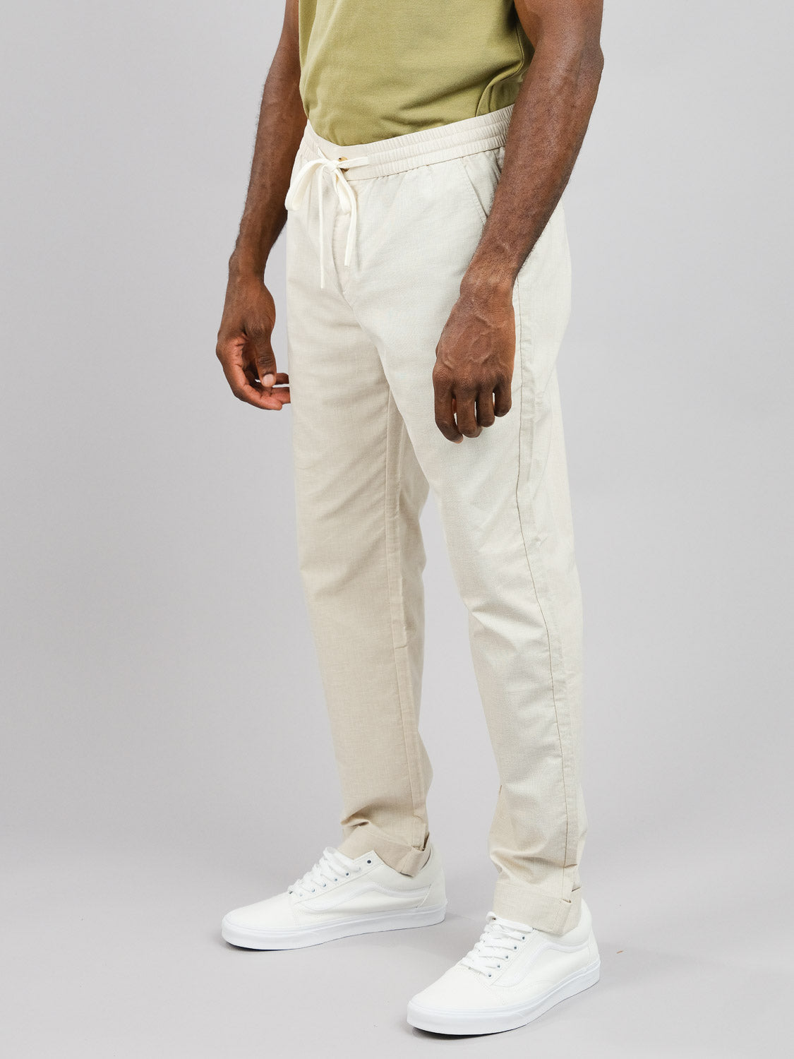 Against a plain background, a person sports stylish comfort in ID Menswears Cruise Lightweight Stretch Pant in light beige, featuring a relaxed fit and drawstring jogger style. Paired with white sneakers and a light green shirt, the ensemble is both chic and cozy.