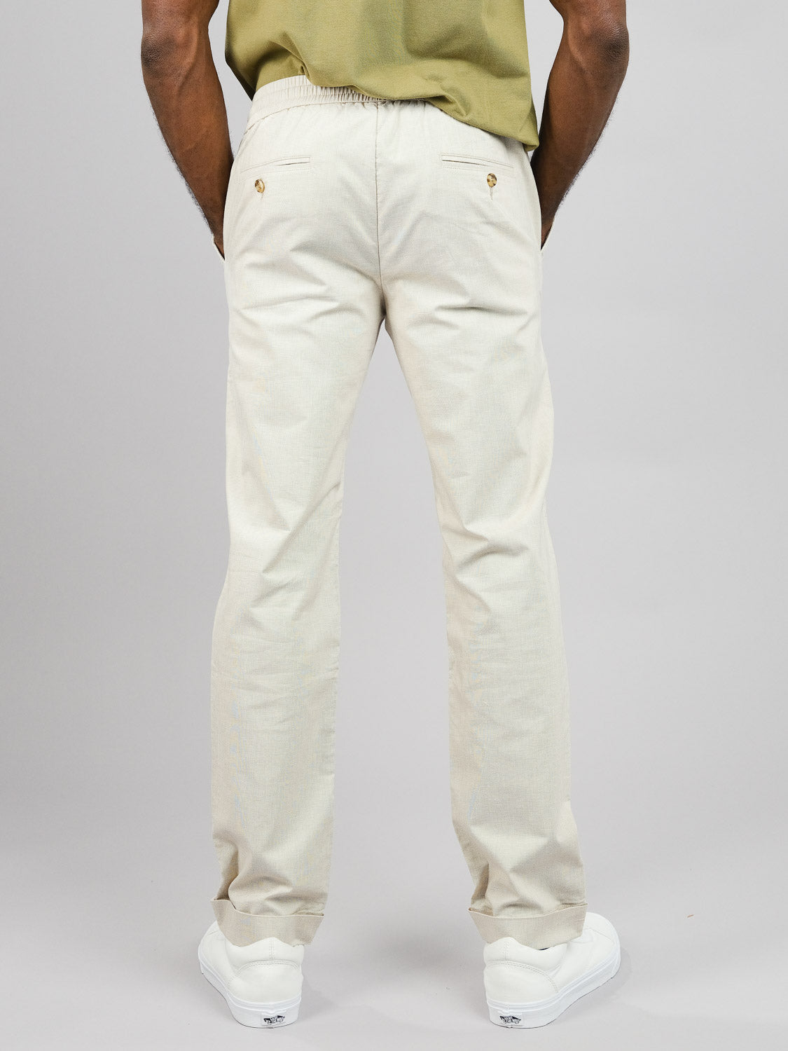 Someone stands with their back to the camera wearing ID Menswears beige Cruise Lightweight Stretch Pant, which includes buttoned back pockets and a drawstring. They pair these pants with a green shirt and white sneakers against a plain gray background.