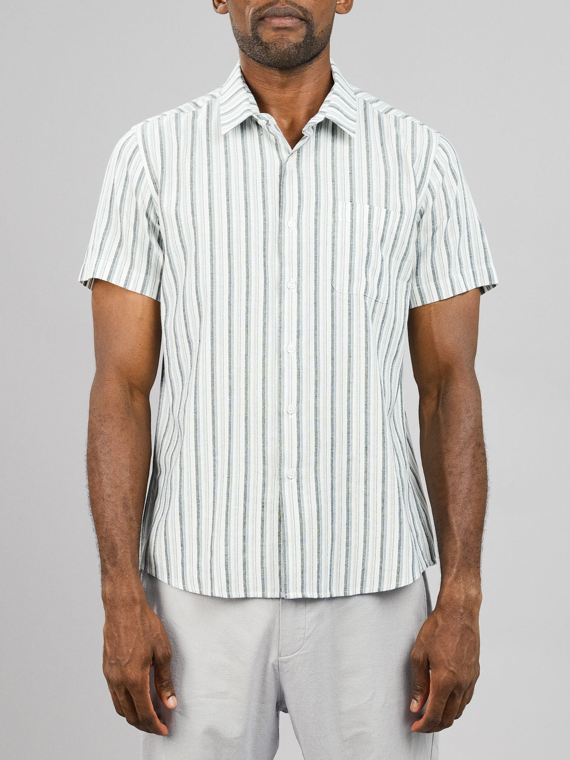 A person stands against a gray background wearing ID Menswears Streak Cotton Shirt, a short-sleeved, lightweight, light blue and white striped button-up shirt, paired with regular fit light gray pants.