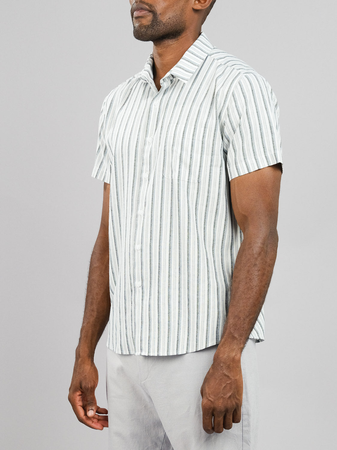 A person models the Streak Cotton Shirt from ID Menswear, featuring a lightweight, light gray and white vertical stripe design and regular fit. The button-up shirt pairs seamlessly with matching light gray pants for an effortlessly relaxed look against a plain background.
