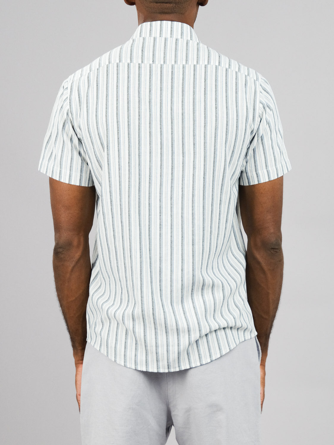 A person wearing an ID Menswear Streak Cotton Shirt with vertical white and gray stripes stands with light gray pants against a plain gray background, creating a cohesive lightweight look.