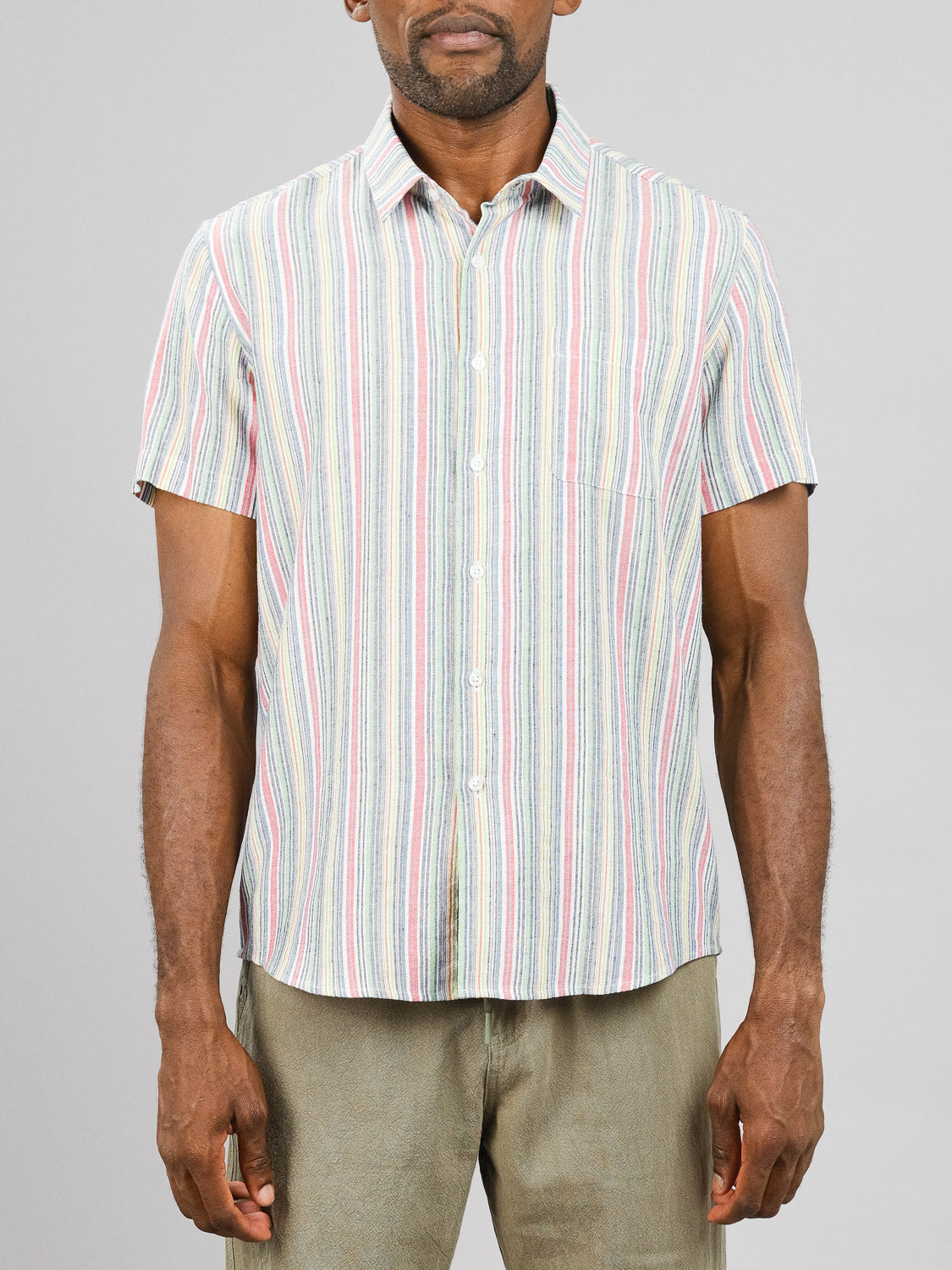 A person wearing the lightweight, regular fit Streak Cotton Shirt by ID Menswear, featuring short sleeves, multicolored vertical stripes, and a left chest pocket stands against a plain background with light-colored pants.