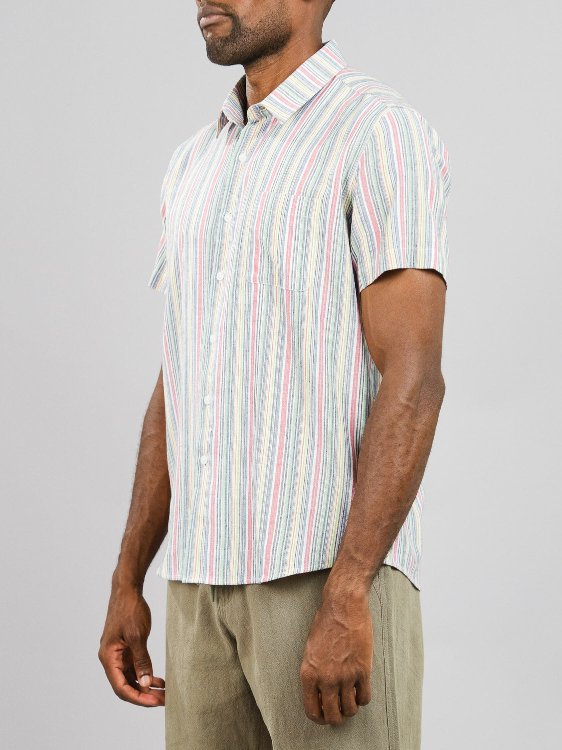 A man wears the Streak Cotton Shirt by ID Menswear, a regular fit lightweight cotton shirt featuring short sleeves and pastel vertical stripes of pink, green, and blue. He pairs it with light olive pants against a plain gray background.