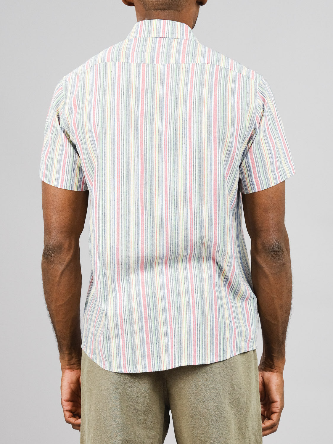 The person showcases the back of the outfit featuring ID Menswears Streak Cotton Shirt—a short-sleeved, lightweight, multicolored striped cotton shirt in a regular fit—paired with beige pants against a light gray backdrop.