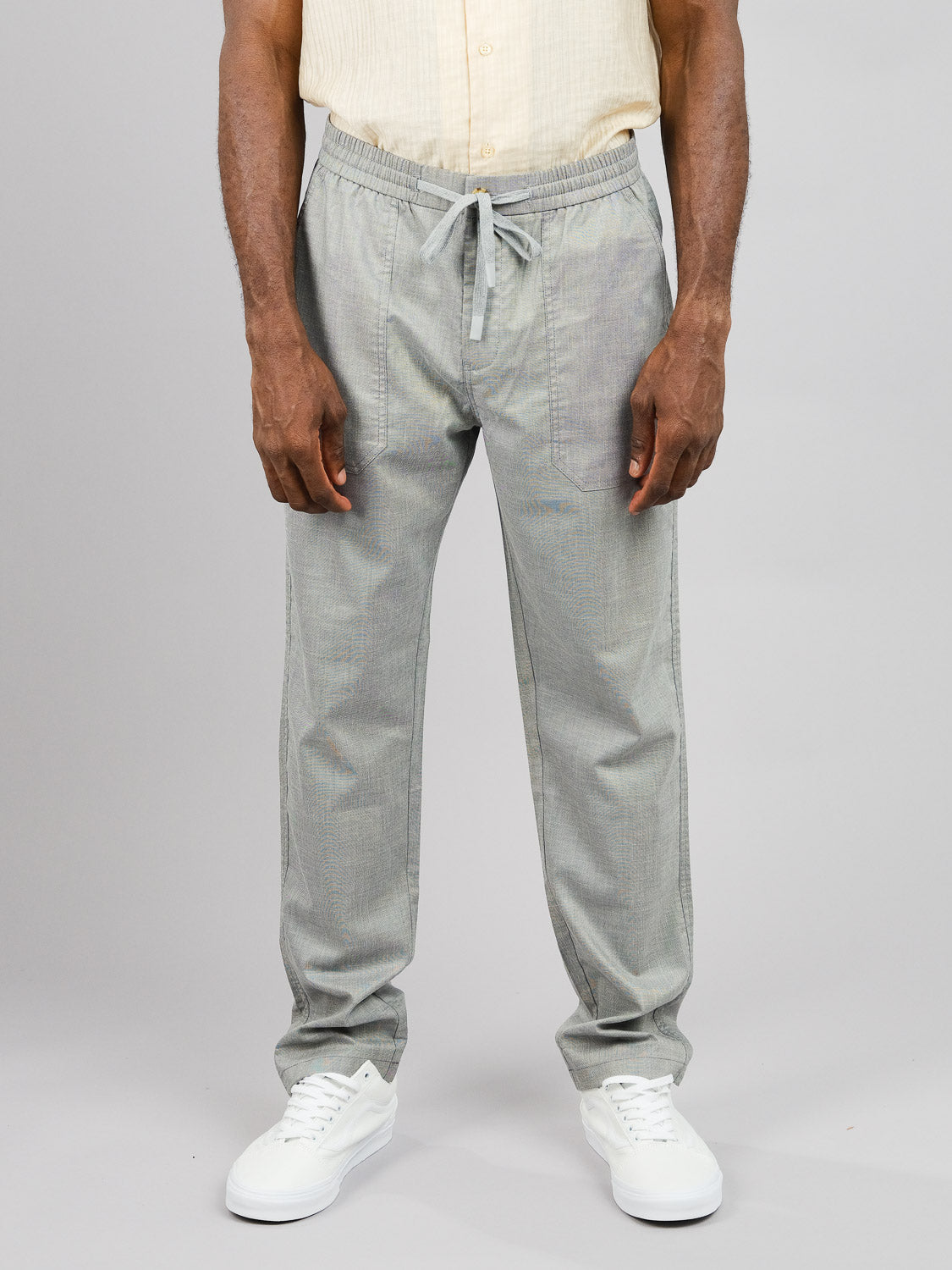 Standing against a gray backdrop, the individual wears ID Menswears Kruger Relaxed Pant with a light yellow shirt and white sneakers. With hands resting at their sides, the ensemble captures warm-weather comfort and an effortlessly casual style.