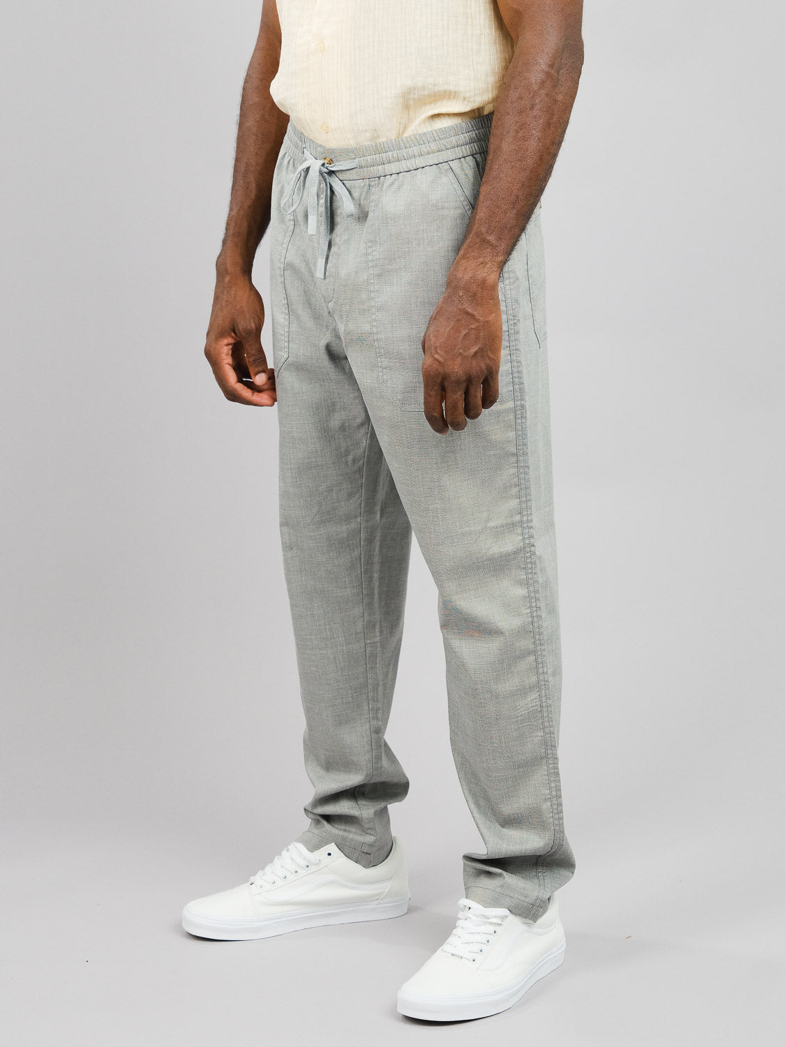 A man dons the Kruger Relaxed Pant by ID Menswear, featuring a relaxed fit and light gray shade with a drawstring waist, paired with a light beige shirt and white sneakers. The warm-weather ensemble pops against the plain gray backdrop.