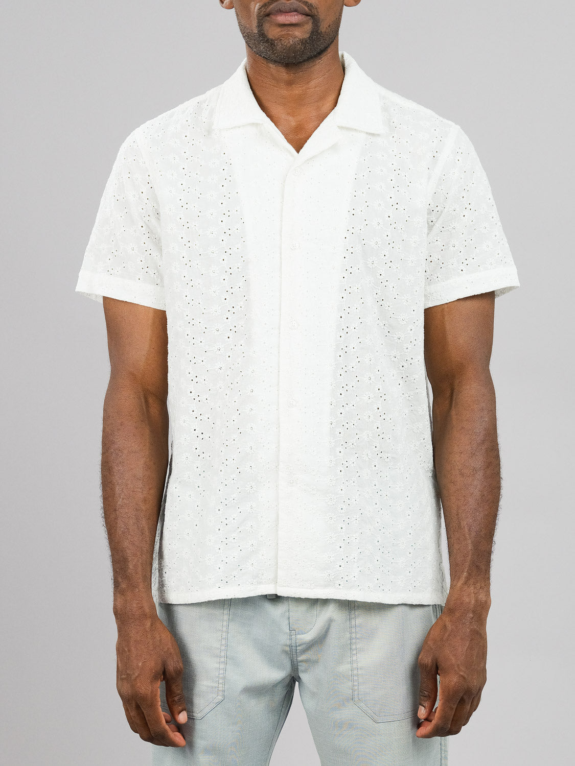 A person models the Anglaise Embroidery Shirt by ID Menswear, featuring a white eyelet pattern against a gray background. Paired with light blue jeans, the casual shirt is in focus while the face is not visible, emphasizing effortless style.