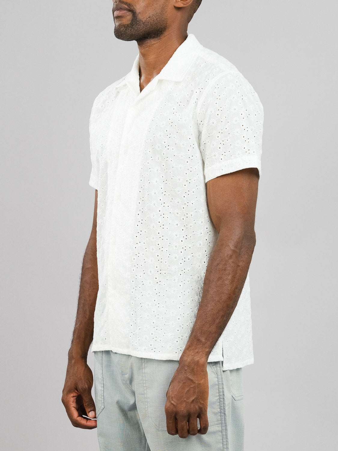 A man in an ID Menswear Anglaise Embroidery Shirt pairs it with light gray pants, standing against a gray background. His head turns slightly left, while his hands rest relaxed by his sides.