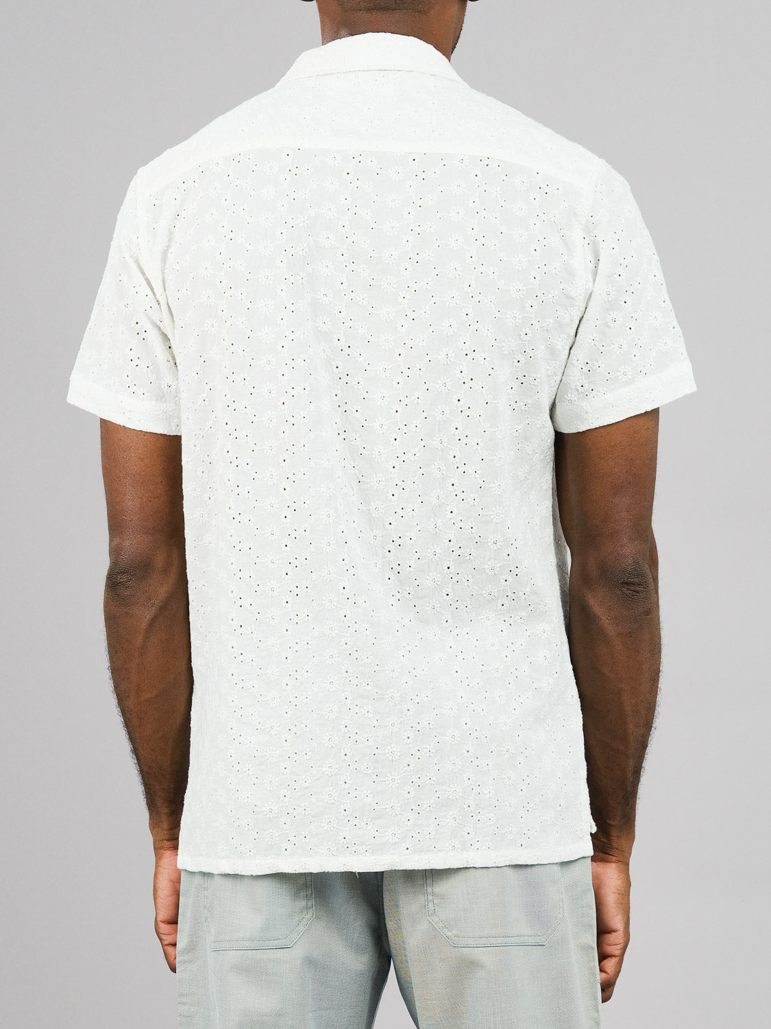 A person wearing the ID Menswear Anglaise Embroidery Shirt, which features a camp collar with a subtle pattern and short sleeves, stands with their back to the camera against a plain gray background. Gray pants complete this casual look.