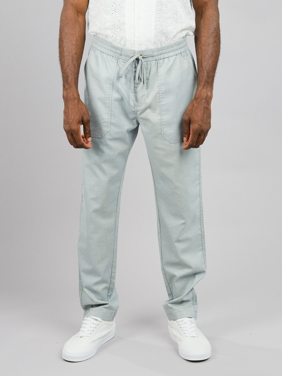 A person is wearing ID Menswear’s Kruger Relaxed Pant in light gray, paired with a white textured shirt and white sneakers. The ensemble offers warm-weather comfort and the individual stands against a plain gray background.