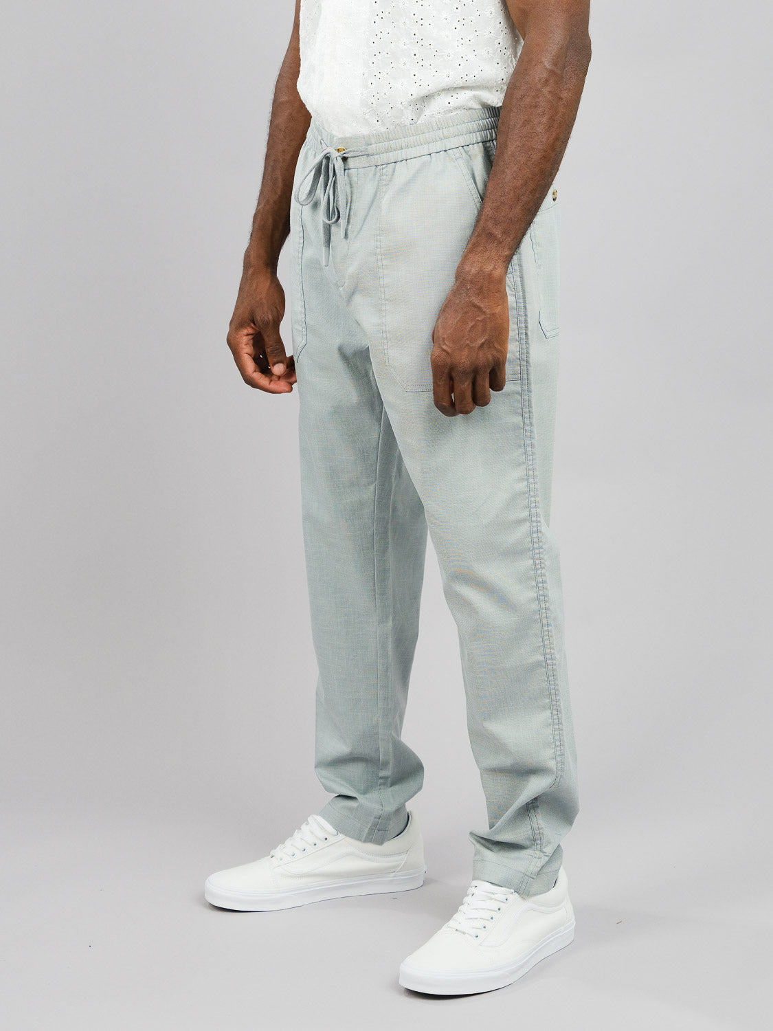 A person embodies warm-weather comfort in ID Menswears Kruger Relaxed Pant, showcasing light gray drawstring pants and white sneakers, paired with a white short-sleeve shirt against a plain gray backdrop. The relaxed fit lets them rest their hands effortlessly by their sides.