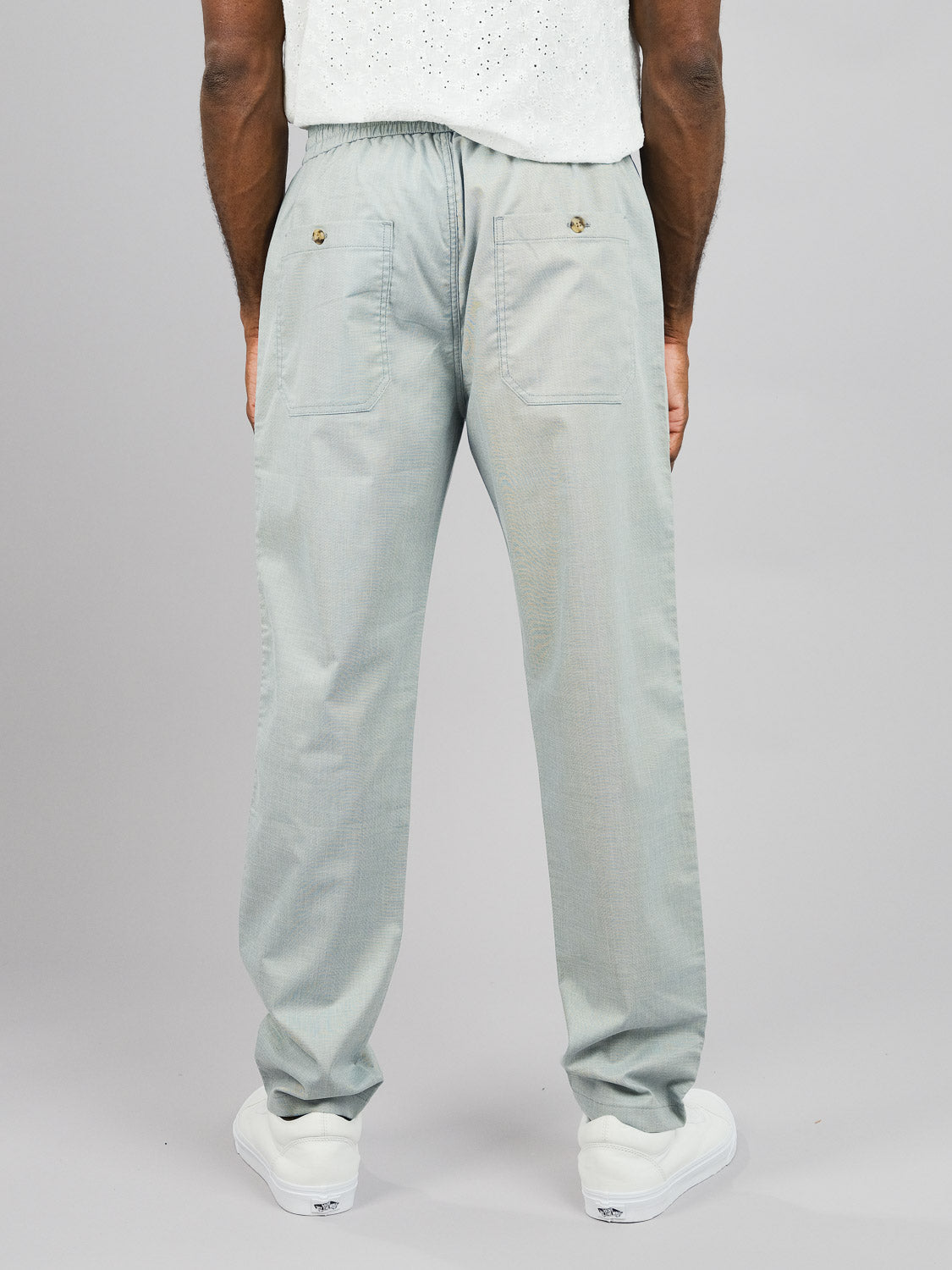 A person is seen from the rear wearing ID Menswears Kruger Relaxed Pant in light gray, featuring an elastic waistband and buttoned back pockets. Paired with a light shirt and white sneakers, this outfit is perfect for warm-weather comfort against a plain gray background.