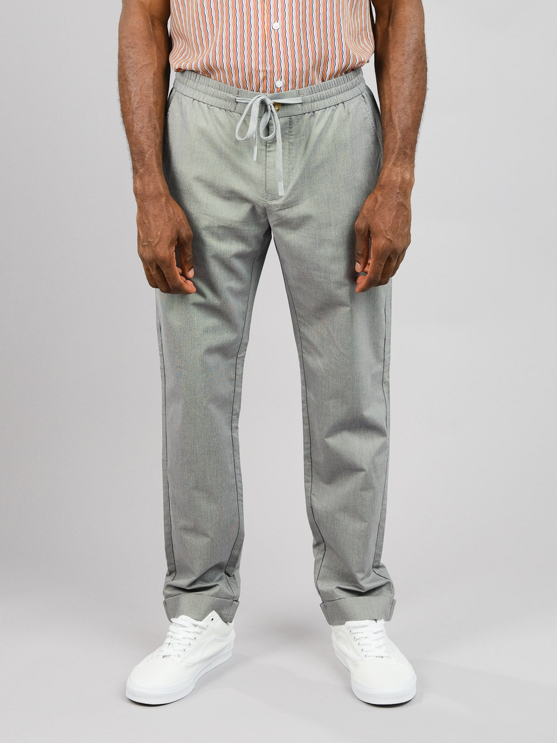 An individual dressed in ID Menswears Cruise Lightweight Stretch Pant in gray and a striped shirt stands against a plain backdrop. The look is finished with white sneakers, drawing attention to the comfort and style of the pants.