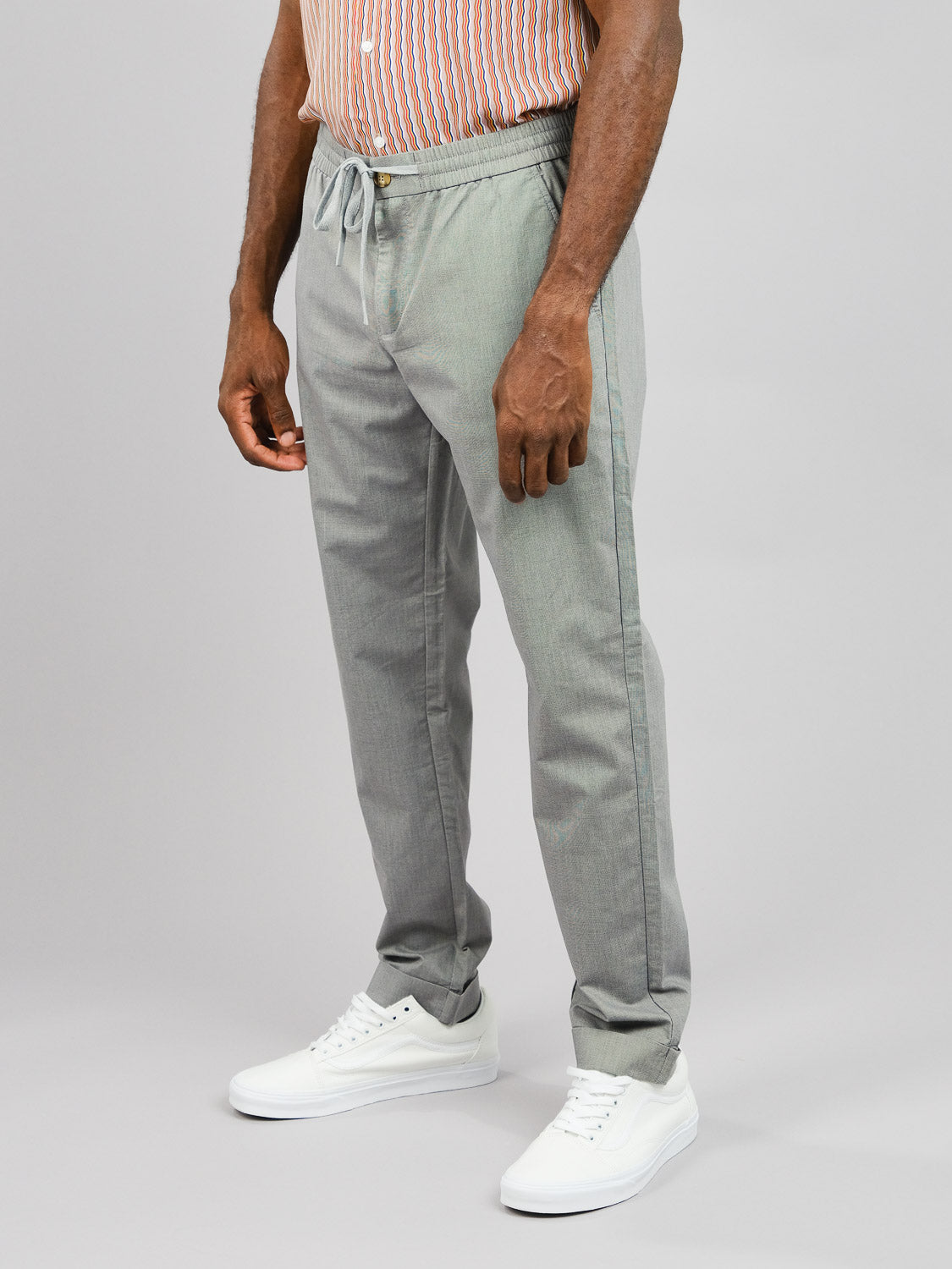 Dressed in ID Menswears Cruise Lightweight Stretch Pant in light gray and a striped shirt, a person stands on a plain gray background wearing white sneakers. Hands relaxed by their sides, they exude effortless style.