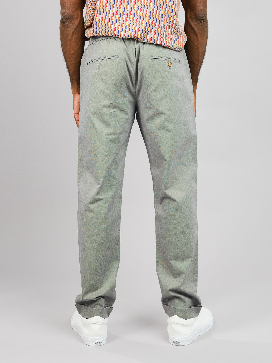 A person stands with their back to the camera, wearing gray ID Menswear Cruise Lightweight Stretch Pants paired with a striped shirt and white sneakers. The pants include pockets and a small decorative detail near one pocket, set against a plain, light background.