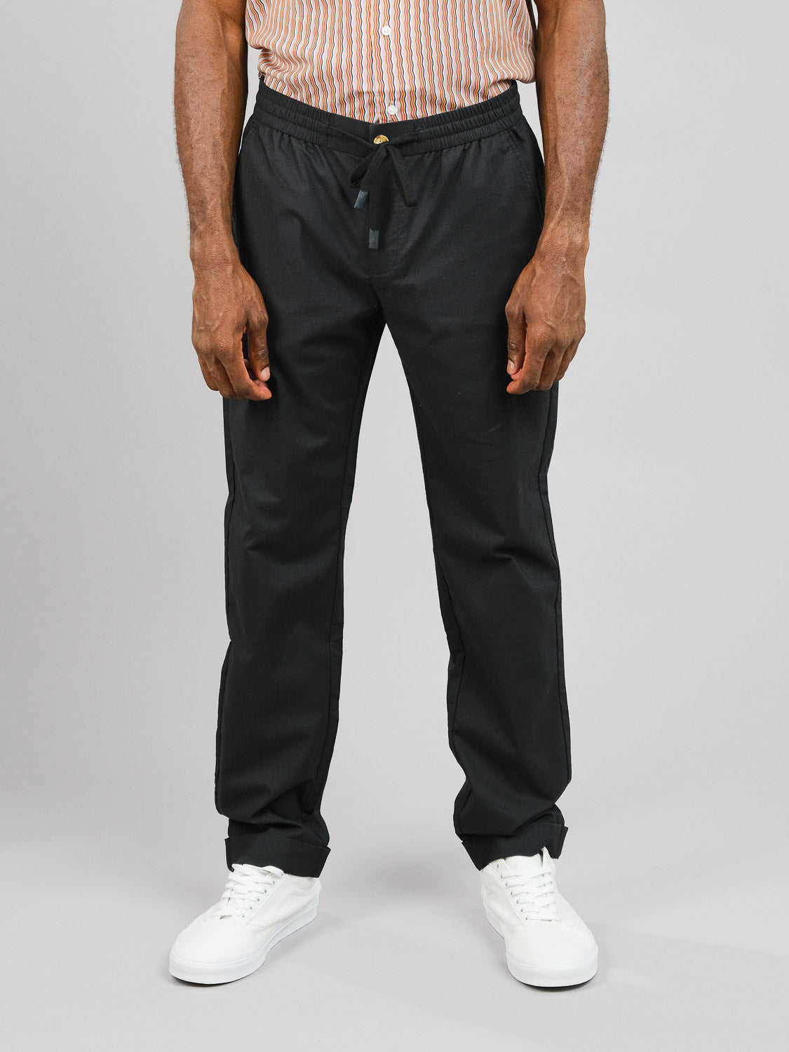 A man exudes casual elegance against a neutral background, wearing the Cruise Lightweight Stretch Pant by ID Menswear and white sneakers. His striped shirt enhances his comfortable yet stylish look.