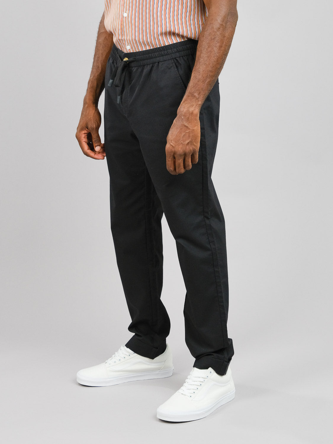 A person in a striped shirt and ID Menswears Cruise Lightweight Stretch Pant stands against a plain background. Complemented by white sneakers, the casual outfit exudes comfort and ease, with only the relaxed lower half visible.