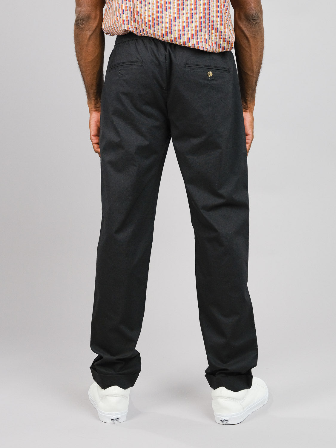 A person stands with their back to the camera, wearing ID Menswears Cruise Lightweight Stretch Pant and white sneakers. The outfit includes a striped shirt, adding to the ensembles airy feel against a plain gray background that enhances the scene’s simple elegance.